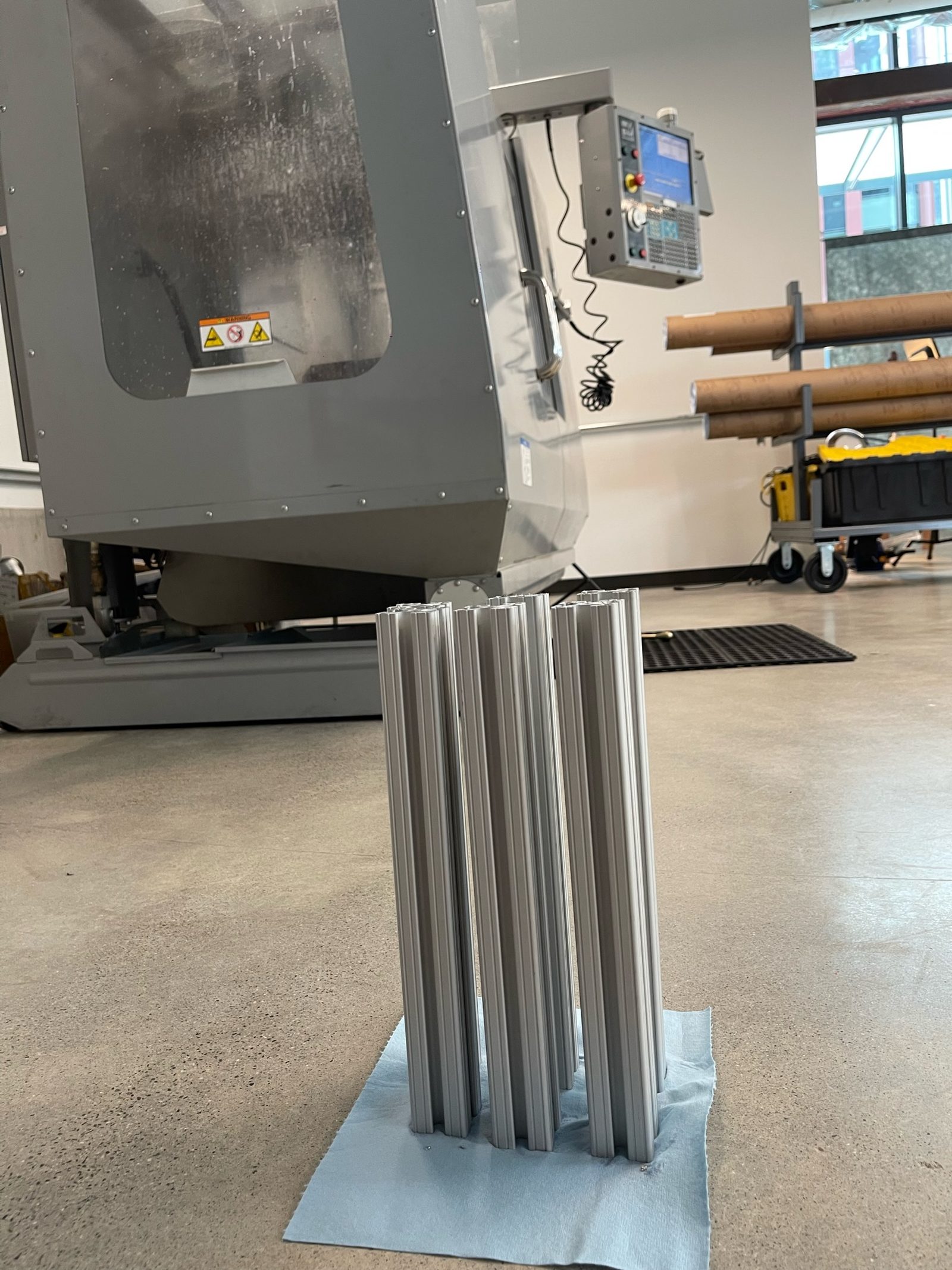 A bundle of cut 8020 aluminium cross-members standing on the workshop floor in front of the Haas Mini Mill, queued for drilling